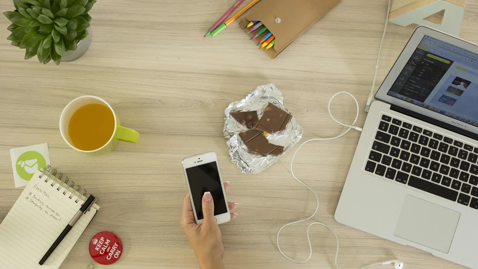 Woman at a table using a smartphone beside a laptop and colored pencils